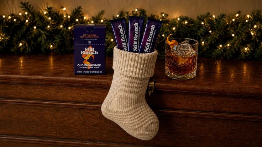 A cocktail stocking stuffer: Still Branch single-serve Old Fashioned packets tucked in a Christmas stocking beside a box and a rocks glass