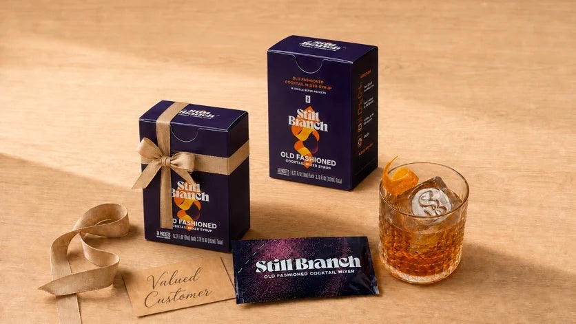 A corporate holiday gift of Still Branch Old Fashioned boxes stacked for clients, with a packet and a finished cocktail over a clear ice cube