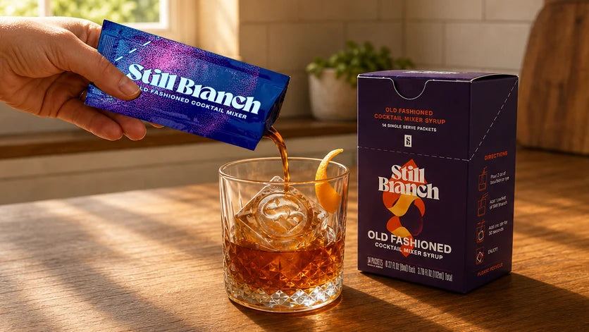 Old Fashioned syrup in a single-serve Still Branch packet poured over a large clear ice cube with bourbon and orange peel