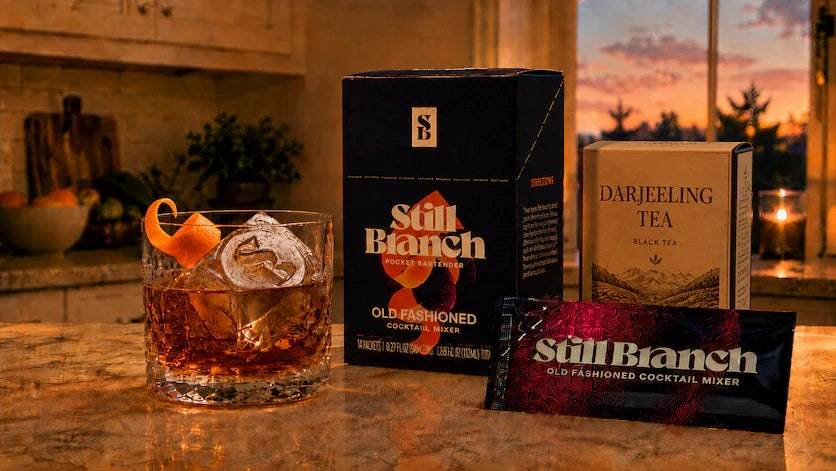 A box and packet of Still Branch with a box of Darjeeling tea next to a Old Fashioned Darjeeling mocktail on a kitchen counter.