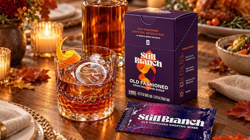 A Thanksgiving Old Fashioned made with a Still Branch packet and bourbon over a clear ice cube, on a table set for the holiday