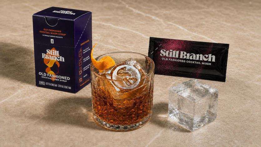Clear ice cube in a Still Branch Old Fashioned built with a single-serve mixer packet and an orange peel next to a clear ice cube on a kitchen counrfer