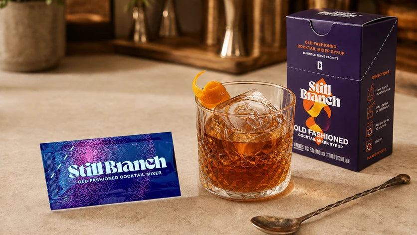 A single-serve cocktail packet from Still Branch poured into a rocks glass with bourbon and a large clear ice cube