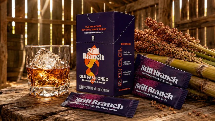 A glass of Old Fashioned cocktail sweetened with sorghum syrup beside a Still Branch box and packet with sorghum cane in a barn