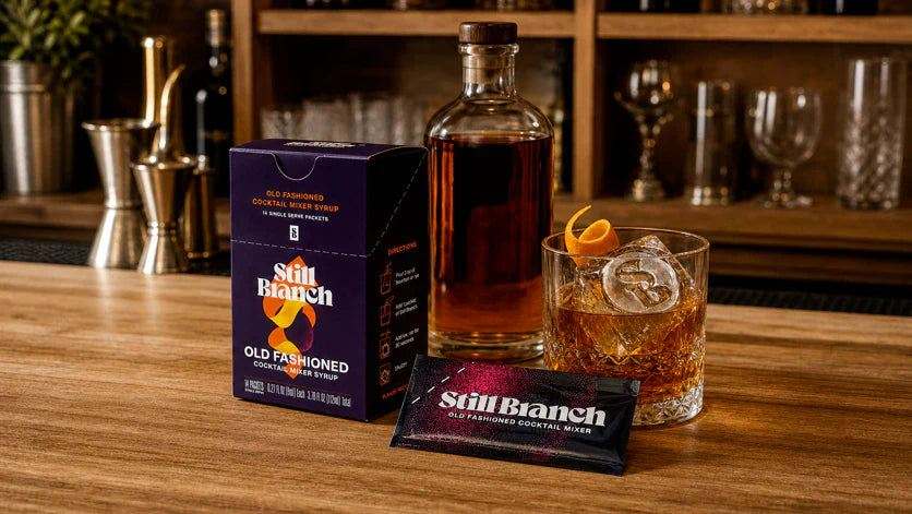 The best Old Fashioned mixer in single-serve Still Branch packets next to a bottle of bourbon and a clear ice cube