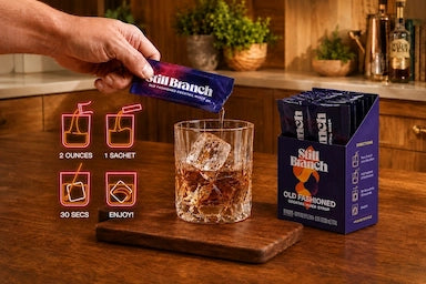 Still Branch Old Fashioned mixer being poured into a glass with ice on a wooden surface.