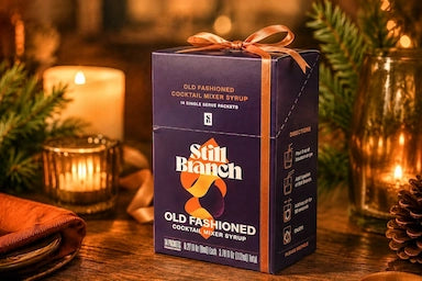 Box of Still Branch Old Fashioned cocktail mixer syrup in a bow on a wooden surface with candles and greenery in the background.