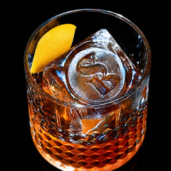 Glass of amber liquid with a lemon wedge and ice cube on a black background